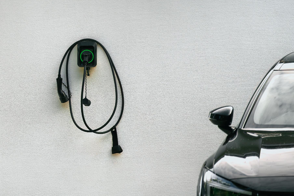 Schwarze Wallbox mit aufgewickeltem Ladekabel an heller Hauswand, daneben Front eines geparkten Elektroautos.